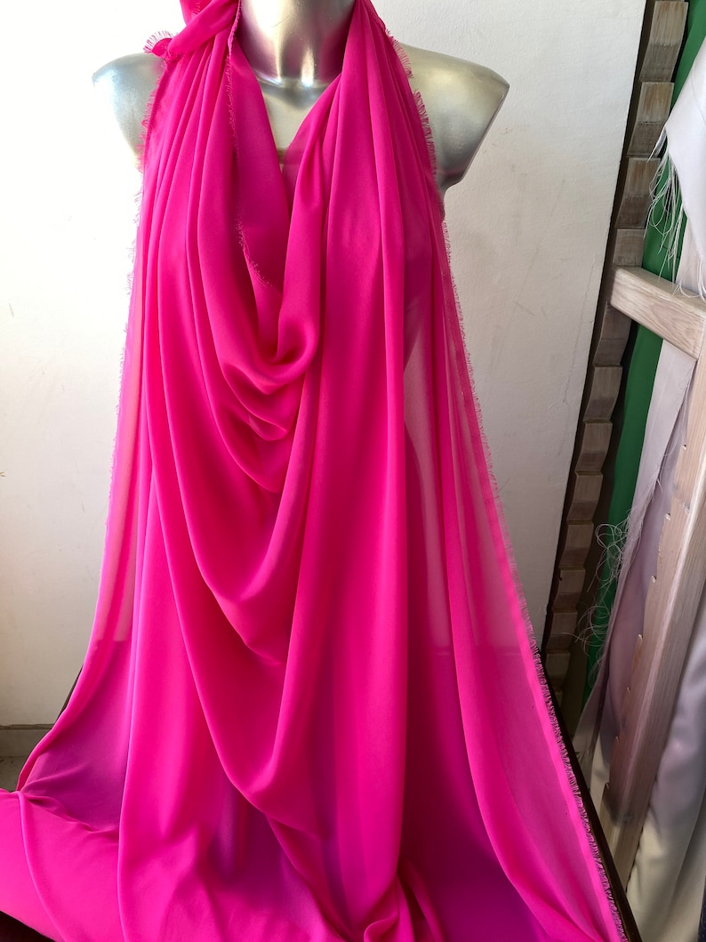 Op de afbeelding: Een helder roze, doorschijnende stof met een rafelige rand. De stof is gedrapeerd over een mannequin.