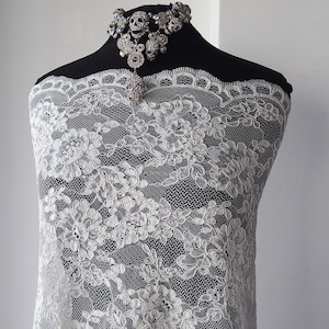May include: White floral lace fabric draped over a black mannequin. The lace has a detailed, repeating pattern of flowers and leaves. A silver skull necklace with rhinestones is displayed at the top.