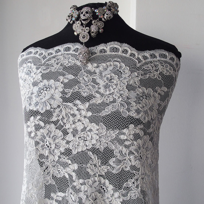 May include: White floral lace fabric draped over a black mannequin. The lace has a detailed, repeating pattern of flowers and leaves. A silver skull necklace with rhinestones is displayed at the top.