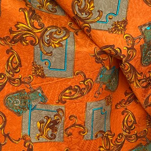 K&ouml;nnte beinhalten: Orangener Stoff mit einem sich wiederholenden Muster aus goldenen Barockmustern und blauen Akzenten. Der Stoff hat eine strukturierte Oberfl&auml;che.