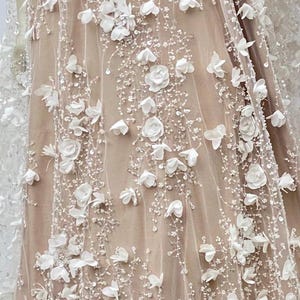 Peut inclure: Gros plan sur un tissu transparent avec des embellissements floraux 3D blancs. Le tissu a un design d&eacute;licat et complexe avec de petites perles et des paillettes.