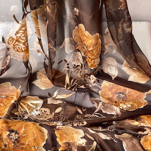 May include: A sheer brown fabric with a floral pattern in shades of brown and gold. The fabric is draped and folded, showing the texture and detail of the print.