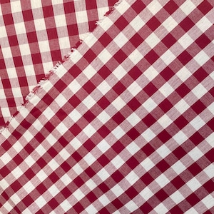 May include: Close-up of red and white gingham fabric. The fabric is cut on a diagonal, with a raw edge visible. The pattern is a classic checkered design, perfect for various crafting projects.