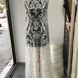 May include: White lace fabric with a floral pattern and beaded accents. The fabric is draped over a dress form.
