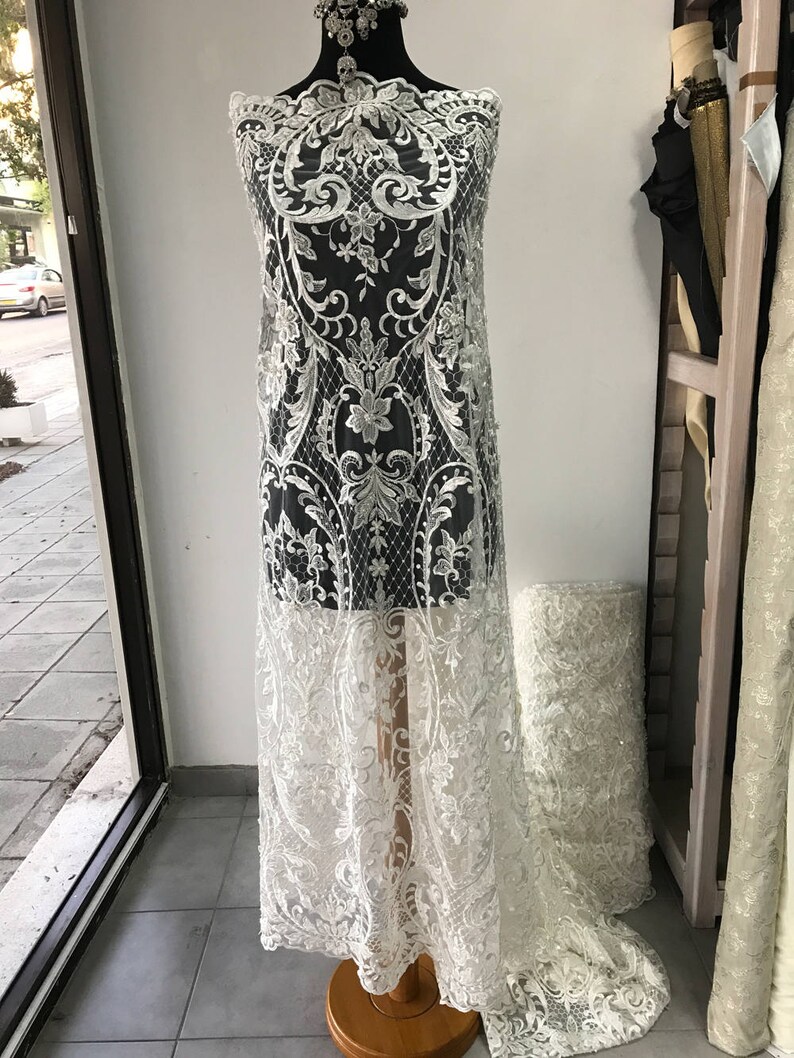 May include: White lace fabric with a floral pattern and beaded accents. The fabric is draped over a dress form.