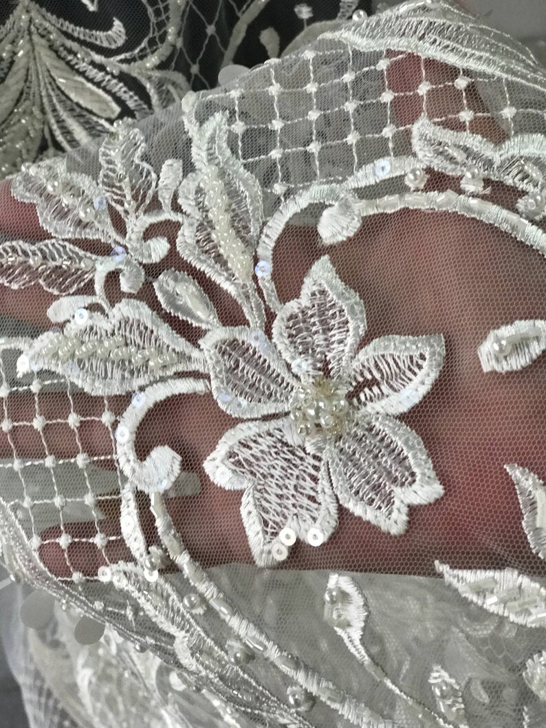 May include: White lace fabric with a floral pattern and pearl embellishments. The lace is delicate and intricate, with a soft, flowing texture.
