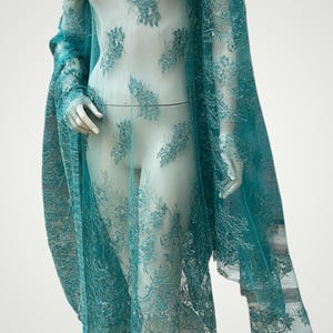 Könnte beinhalten: Ein tealfarbenes Spitzenkleid auf einer Schaufensterpuppe. Das Kleid hat detaillierte Blumenmuster und ein fließendes Design, einschließlich eines Capes. Der Stoff ist zart und halbtransparent, mit langen Ärmeln und einem knielangen Rock.