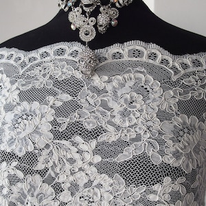 May include: Close-up of white lace fabric with a floral design. The lace is draped over a black form, and a silver skull pendant necklace with rhinestones is visible. The lace has a delicate, open weave.