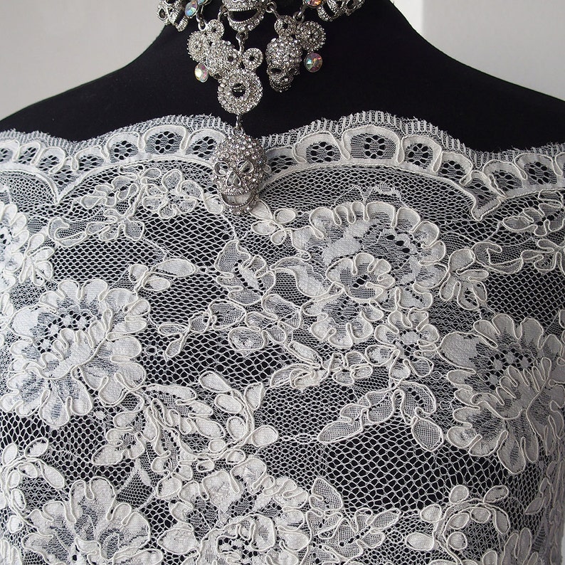 May include: Close-up of white lace fabric with a floral design. The lace is draped over a black form, and a silver skull pendant necklace with rhinestones is visible. The lace has a delicate, open weave.