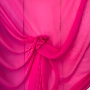 Op de afbeelding: Een close-up van een helder roze, doorschijnende stof, gevouwen en gedrapeerd. De stof is zacht en vloeibaar, met een delicate textuur.