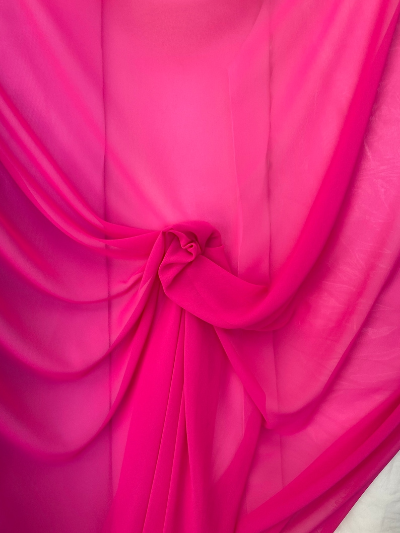 Op de afbeelding: Een close-up van een helder roze, doorschijnende stof, gevouwen en gedrapeerd. De stof is zacht en vloeibaar, met een delicate textuur.