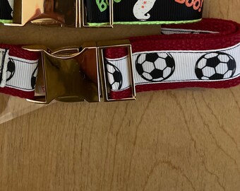 Peut inclure: Deux colliers pour chiens avec des boucles en métal doré. L'un est noir avec une bordure vert fluo et présente un fantôme et le mot "Boo!". L'autre est rouge avec des graphiques de ballon de football.