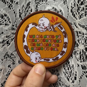 May include: Circular embroidered patch with a brown edge and a mustard yellow centre. The patch features a cartoon character with a knife and the text "WE ALL NEED TO WORK TOGETHER OR WE ARE ALL GOING TO DIE."