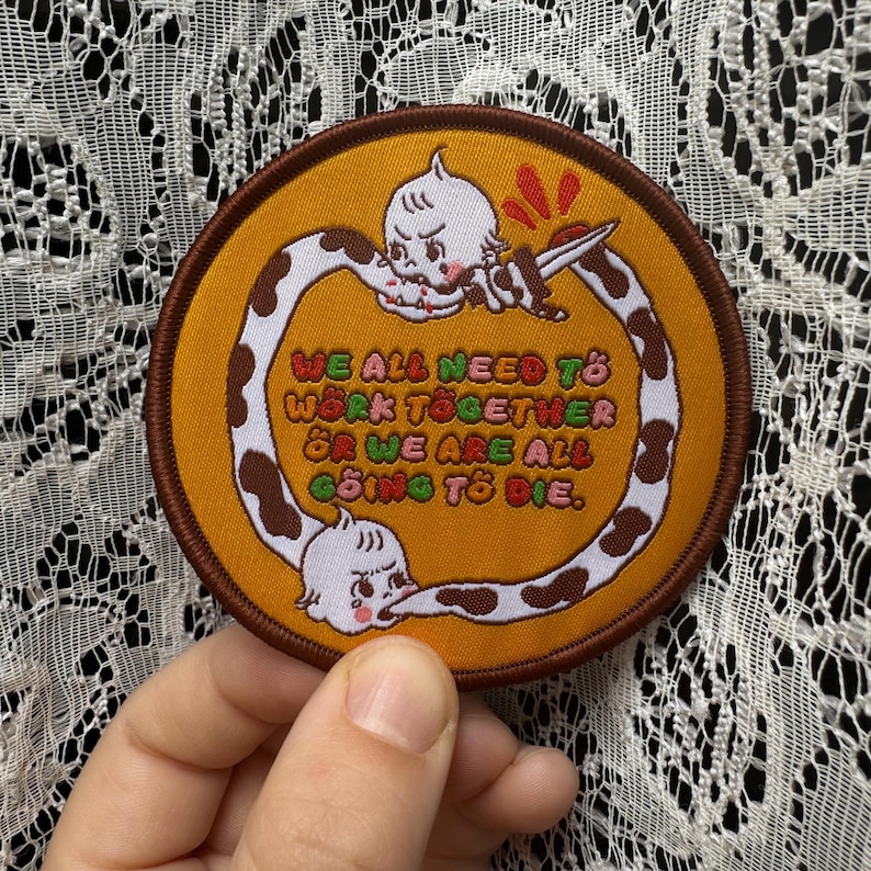 May include: Round embroidered patch with a brown border and a mustard yellow center. The patch features a cartoon character with a knife and the text "WE ALL NEED TO WORK TOGETHER OR WE ARE ALL GOING TO DIE."