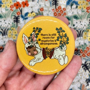 May include: A yellow button featuring a cartoon bunny wearing a corset and a floral crown. The text on the button reads "There is still room for mysteries & strangeness."