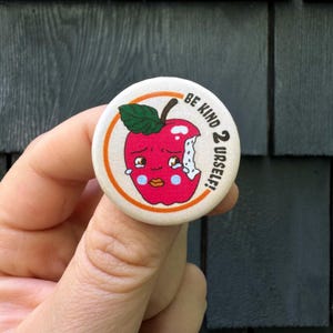 May include: A round button with a white background and an orange border. The button features a cartoon illustration of a red apple with a bite taken out of it. The apple has a face with tearful eyes and a pink mouth. The text "BE KIND 2 URSELEF!" is written in black around the apple.