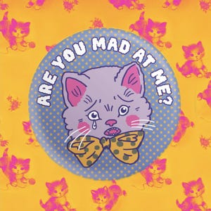 Op de afbeelding: Een blauwe en witte knop met een cartoonkat die een gele en bruine strik draagt. De kat heeft een traan uit zijn oog en de tekst "Are you mad at me?" staat rond de kat.