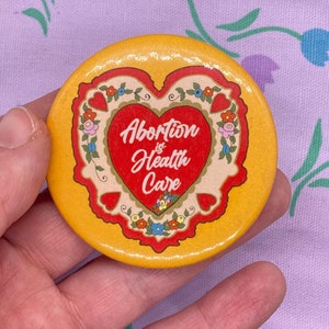 May include: A yellow button featuring a red heart design. The heart has a white centre with the text "Abortion is Health Care" in red lettering. The heart is surrounded by a floral border.