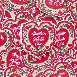 May include: A stack of red heart-shaped stickers with a white border and floral designs. The stickers have the text "Abortion is Health Care" in white lettering.