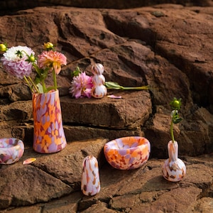 May include: A selection of decorative glass vases and bowls with a speckled orange, purple, and white design. One vase contains flowers. The items are arranged on a brown, textured rock surface.