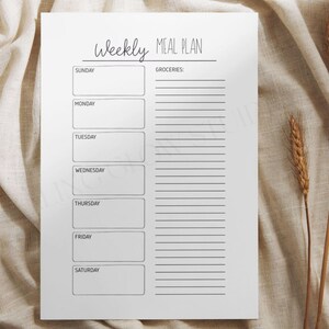 Könnte beinhalten: Ein weißes Wochenessensplan-Blatt mit den Worten "Weekly Meal Plan" oben. Das Blatt hat Kästchen für jeden Wochentag und einen Abschnitt für Lebensmittel. Das Blatt liegt auf einer beigen Oberfläche.