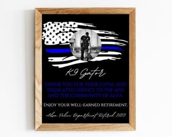 May include: A framed print with a black background and a blue and white American flag with a thin blue line. The flag has a circle in the center with a photo of a police officer. The text "K9 Gafor" is below the flag. The text "Thank you for your loyal and dedicated service to the APD and the community of Alva. Enjoy your well-earned retirement. Alva Police Department Retired 2023" is below the photo.