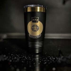 May include: Black insulated tumbler with a gold-colored rim and lid. The tumbler features a gold emblem with the text "772 OKLAHOMA HIGHWAY PATROL TROOPER". The tumbler has a textured surface and a clear lid.