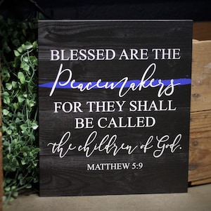 May include: A black wooden sign with white text that reads "Blessed are the Peacemakers for they shall be called the children of God. Matthew 5:9". A blue line is painted across the sign.