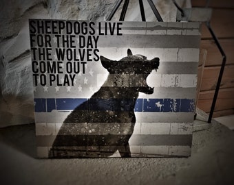 Puede incluir: Una imagen en blanco y negro de un perro con la boca abierta, silueteado sobre un fondo de bandera estadounidense desgastada. El texto "SHEEPDOGS LIVE FOR THE DAY THE WOLVES COME OUT TO PLAY" está impreso en la bandera.