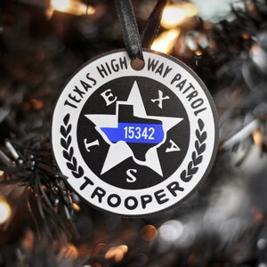May include: A black and white round ornament with the text "Texas Highway Patrol Trooper" and a blue and white outline of the state of Texas with the number "15342" inside.