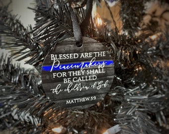 May include: A round wooden ornament with a black and white woodgrain pattern. It features a blue ribbon with a thin white line, and the text "BLESSED ARE THE Peacemakers FOR THEY SHALL BE CALLED the children of God. MATTHEW 5:9".