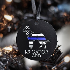 May include: A black round Christmas ornament with a white silhouette of a German Shepherd dog. The dog is depicted with an American flag pattern and a blue line across its chest. The text "K9 GATOR APD" is printed below the dog.
