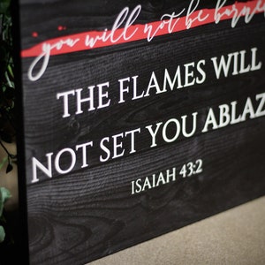 Puede incluir: Un letrero de madera negro con texto blanco que dice "THE FLAMES WILL NOT SET YOU ABLAZE" y "ISAIAH 43:2". La parte superior del letrero tiene el texto "you will not be burned" en escritura blanca con un acento rojo. El letrero tiene una textura r&uacute;stica de vetas de madera.