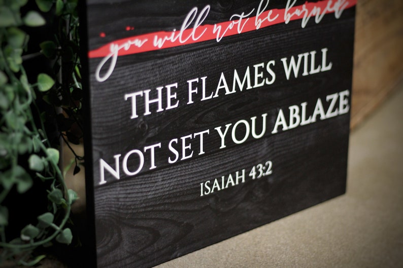 Puede incluir: Un letrero de madera negro con texto blanco que dice "THE FLAMES WILL NOT SET YOU ABLAZE" y "ISAIAH 43:2". La parte superior del letrero tiene el texto "you will not be burned" en escritura blanca con un acento rojo. El letrero tiene una textura r&uacute;stica de vetas de madera.