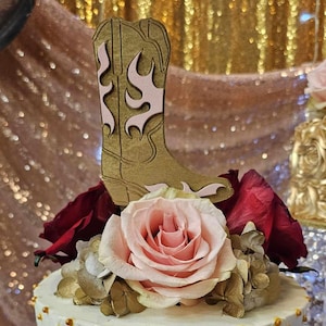 May include: A white cake with gold accents and a quilted design. The cake is decorated with a pink rose, burgundy roses, and a wooden cutout of a cowboy boot.
