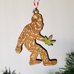May include: Wooden Christmas ornament featuring a cartoon Bigfoot holding a green marijuana leaf. The ornament is brown and has a string for hanging.