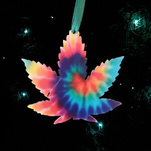 May include: A tie-dye patterned leaf ornament in shades of pink, blue, and yellow. The ornament is hanging from a green ribbon against a black background.