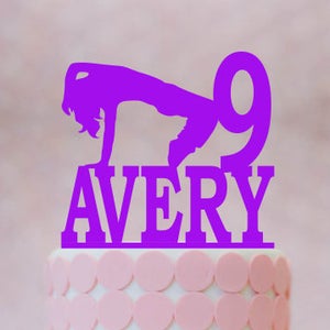 May include: Purple cake topper with a silhouette of a girl doing a handstand and the number 9. The topper also includes the name Avery.