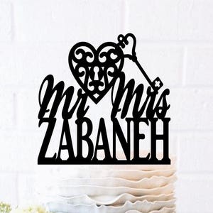 May include: A black cake topper with the words "Mr & Mrs ZABANEH" in a script font. The topper features a heart with a key design. The cake is decorated with blue and white ruffles and adorned with flowers.