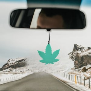 May include: A teal green marijuana leaf car air freshener hanging from the rearview mirror of a car driving on a snowy road.