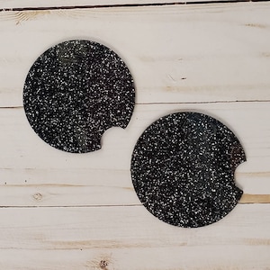Pu&ograve; includere: Due sottobicchieri rotondi neri con una finitura glitterata bianca. I sottobicchieri hanno un'apertura per una tazza.