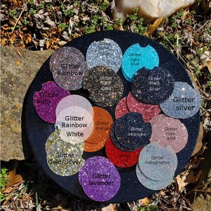 Pu&ograve; includere: Una variet&agrave; di sottobicchieri rotondi ricoperti di glitter in diversi colori, tra cui rubino, arcobaleno, bianco, arancione, mezzanotte, oro rosa, oro e argento, lavanda, rosso, olografico, argento spesso, blu chiaro, oro nero e argento nero. I sottobicchieri sono disposti in un cerchio su una superficie nera.