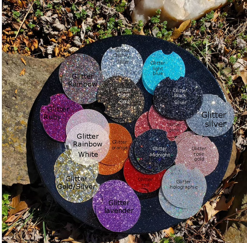 Pu&ograve; includere: Una variet&agrave; di sottobicchieri rotondi ricoperti di glitter in diversi colori, tra cui rubino, arcobaleno, bianco, arancione, mezzanotte, oro rosa, oro e argento, lavanda, rosso, olografico, argento spesso, blu chiaro, oro nero e argento nero. I sottobicchieri sono disposti in un cerchio su una superficie nera.