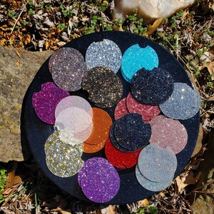 Pu&ograve; includere: Un set di 15 sottobicchieri rotondi e scintillanti in vari colori, tra cui oro, argento, rosa, blu, nero e viola. I sottobicchieri sono disposti in cerchio su una superficie nera.