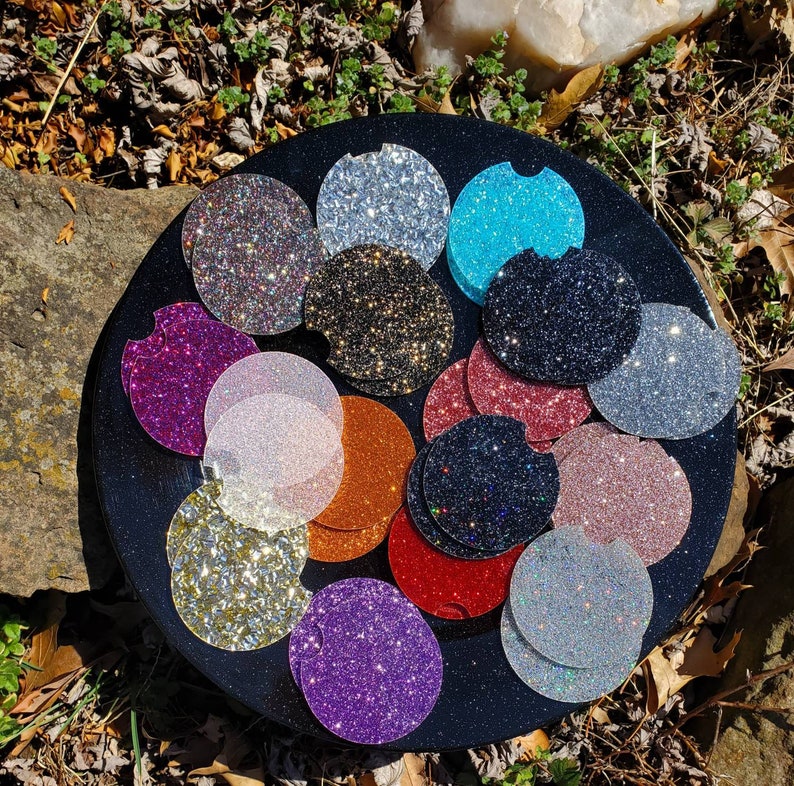 Pu&ograve; includere: Un set di 15 sottobicchieri rotondi e scintillanti in vari colori, tra cui oro, argento, rosa, blu, nero e viola. I sottobicchieri sono disposti in cerchio su una superficie nera.