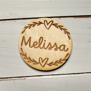 May include: A round wooden ornament with a leafy wreath and two hearts surrounding the name "Melissa".