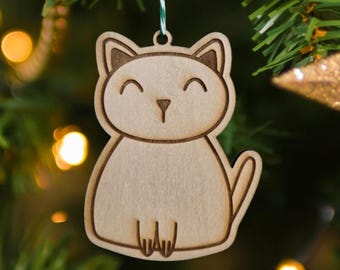 Puede incluir: Adorno navideño de madera con forma de gato. El adorno es de color marrón claro con una cara sonriente y una pequeña cola. Cuelga de una cuerda verde y blanca sobre un fondo borroso de árbol de Navidad.