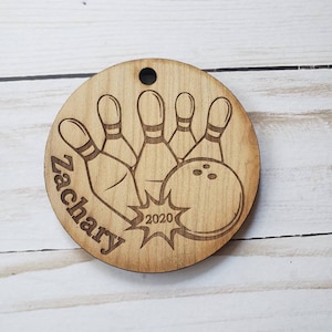 May include: A wooden round ornament with a bowling ball and pins design. The ornament is engraved with the name "Zachary" and the year "2020".