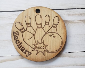 May include: A wooden round ornament with a bowling ball and pins design. The ornament is engraved with the name "Zachary" and the year "2020".