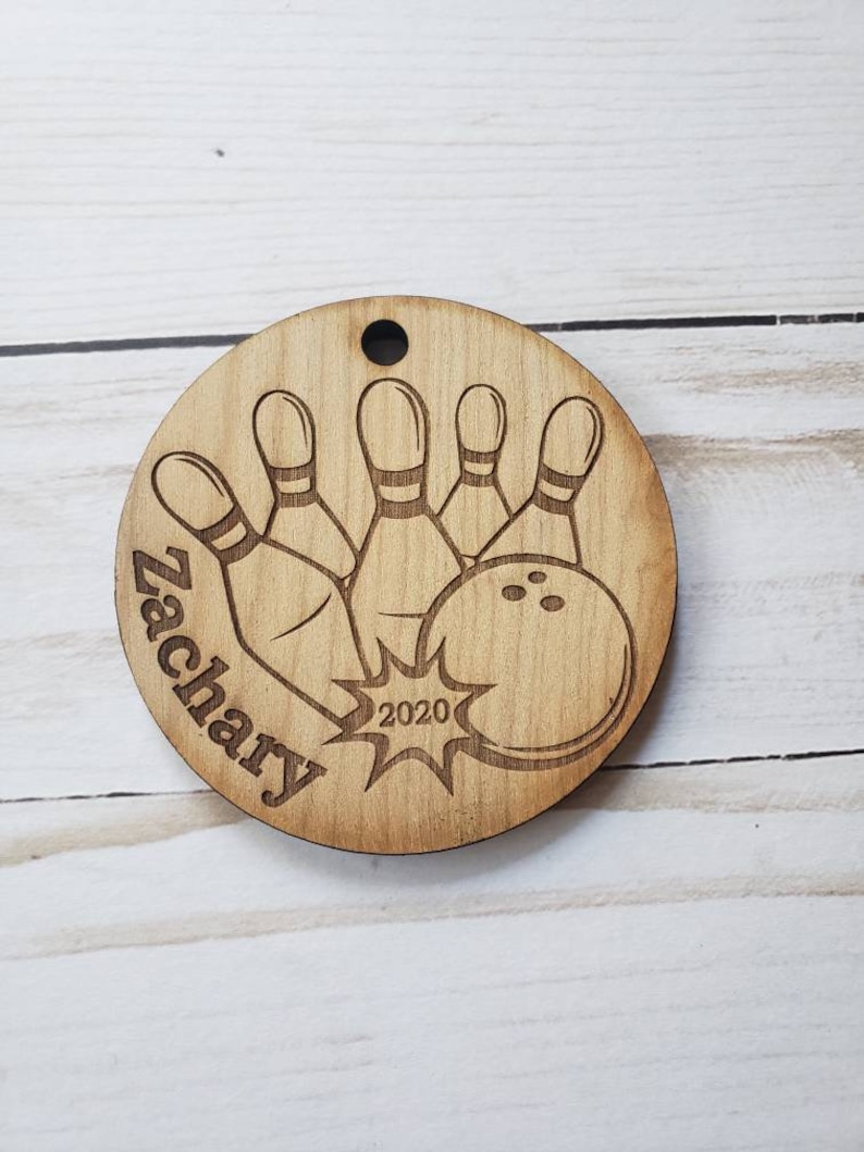 May include: A wooden round ornament with a bowling ball and pins design. The ornament is engraved with the name "Zachary" and the year "2020".